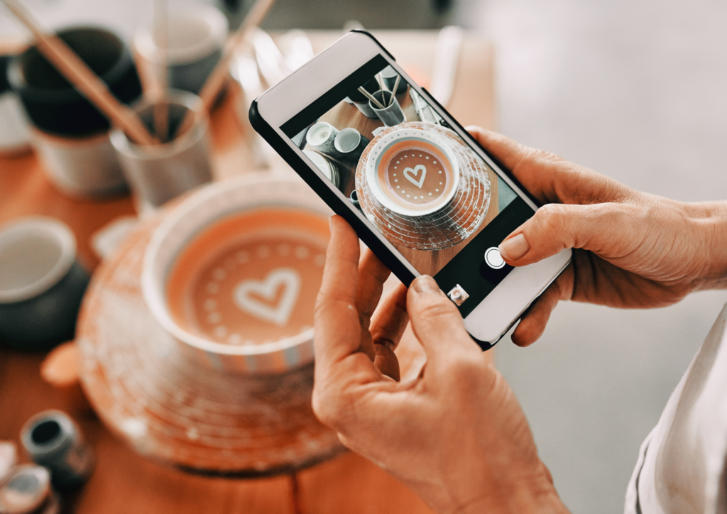 Hands holding a smartphone, taking a photo of a ceramic bowl with a heart design, capturing the intricate details and warm tones of the pottery, while brushes and cups are visible in the blurred background.